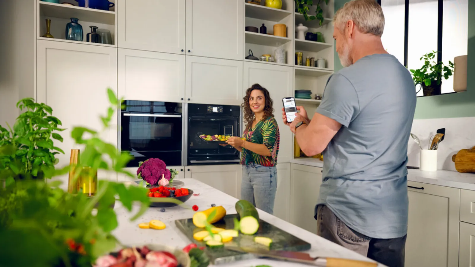 A man and woman stand in a kitchen, holding a phone and discussing something with smiles on their faces.  
