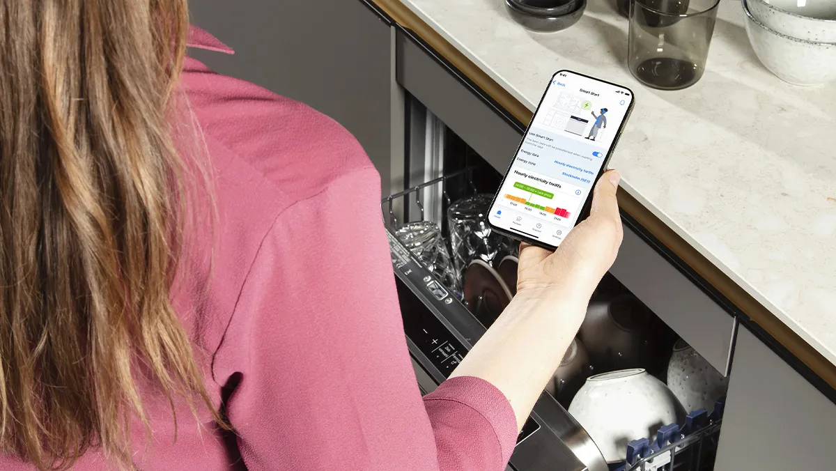 Blick über die Schulter einer Frau, die ein Smartphone mit der Home Connect App über einen geöffneten Geschirrspüler hält, um Verbrauchsdaten abzulesen.