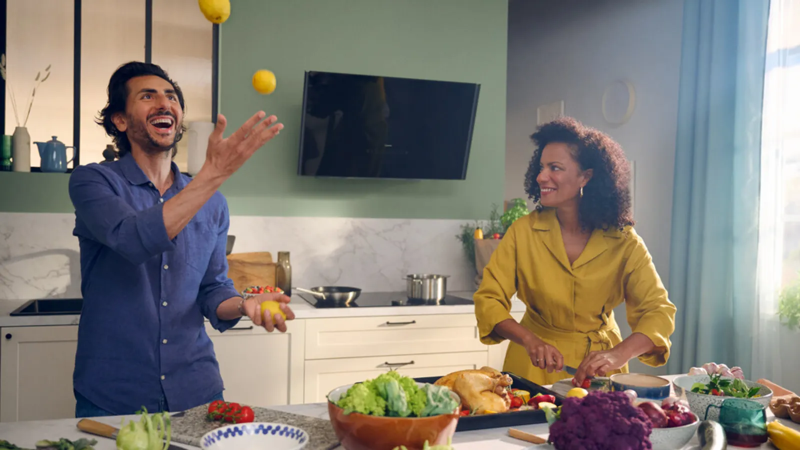 A man and woman in a kitchen surrounded by fruit and vegetables.
