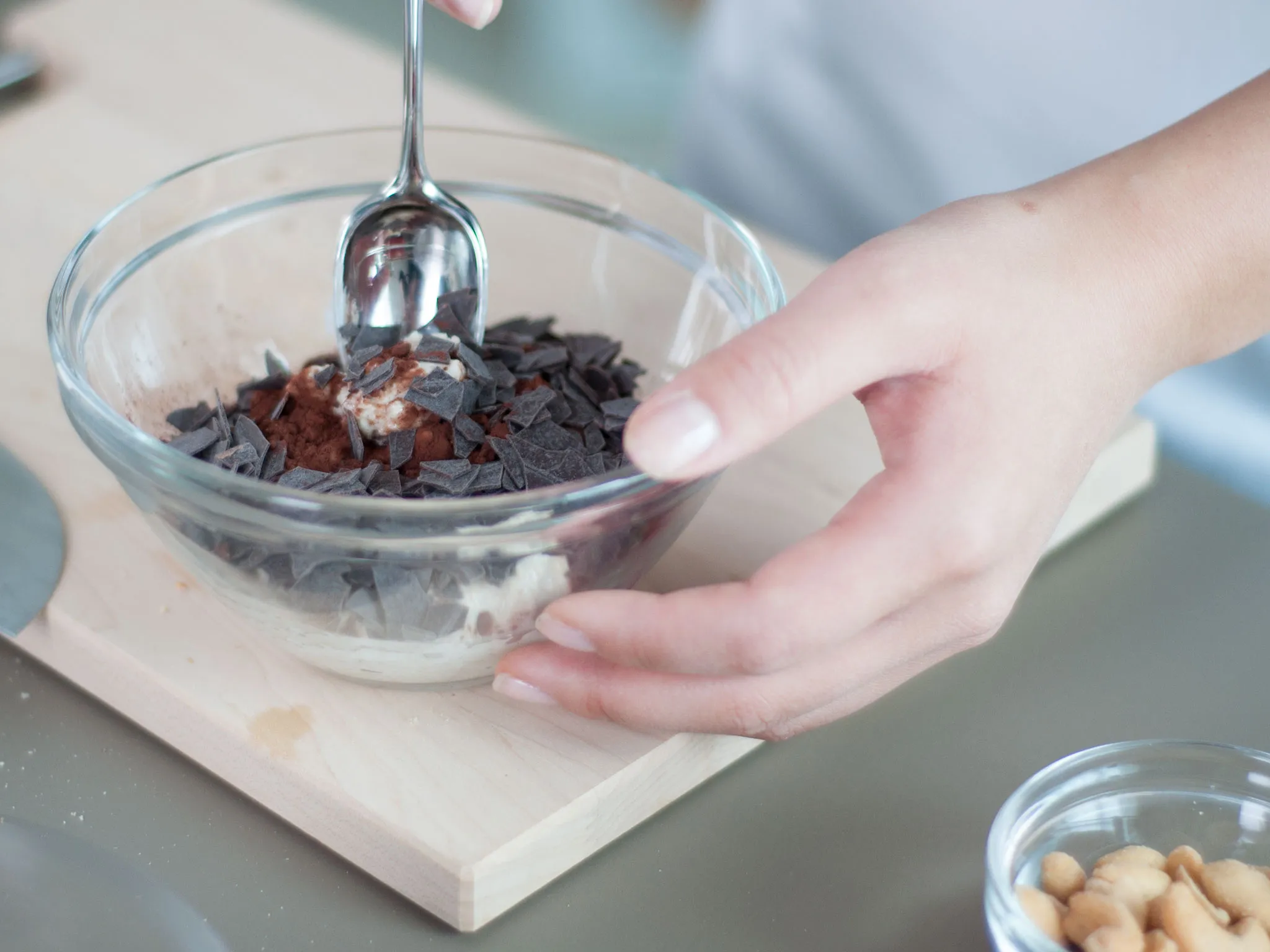 Hands scoop a spoonful from a glass bowl of creamy dessert topped with dark chocolate shavings; a small bowl of peanuts sits nearby.