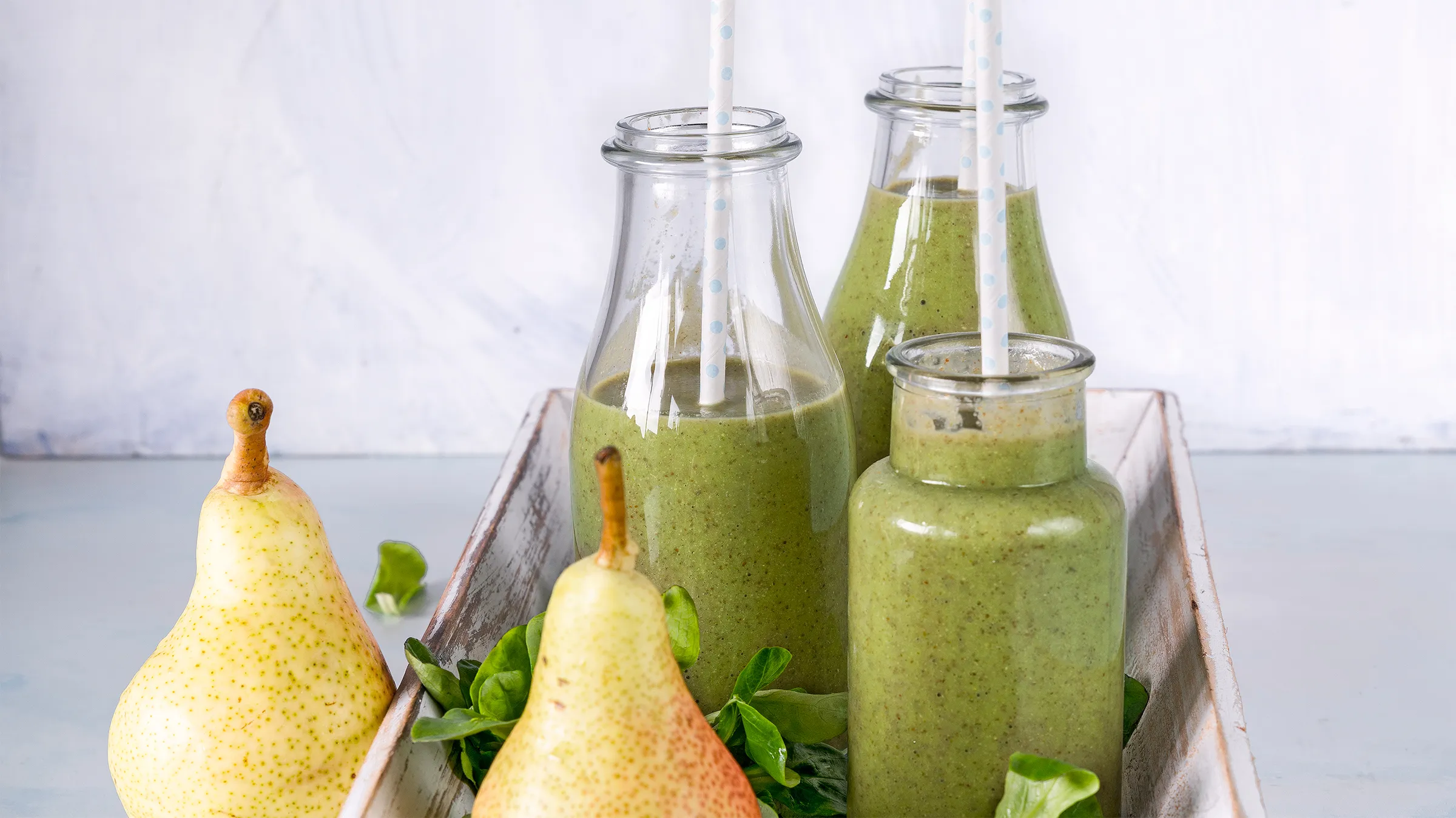 Patru sticle cu smoothie verde, fiecare cu câte un pai, aşezate pe o tavă albă, alături de două pere şi frunze verzi.