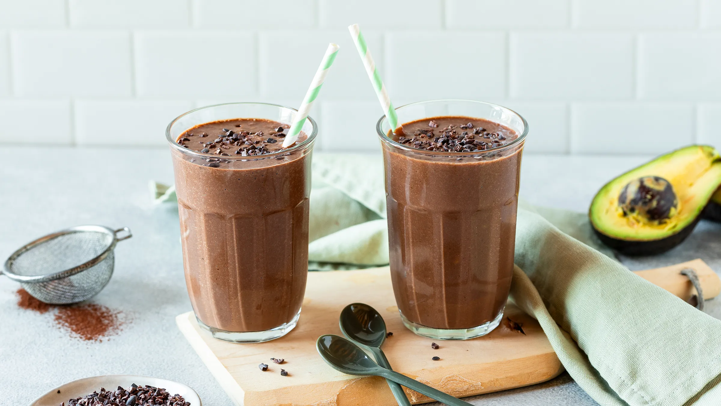 Două pahare de shake de ciocolată cu avocado, cu miezuri de cacao pe o tavă de culoare închisă, cu o lingură de miezuri de cacao alături.