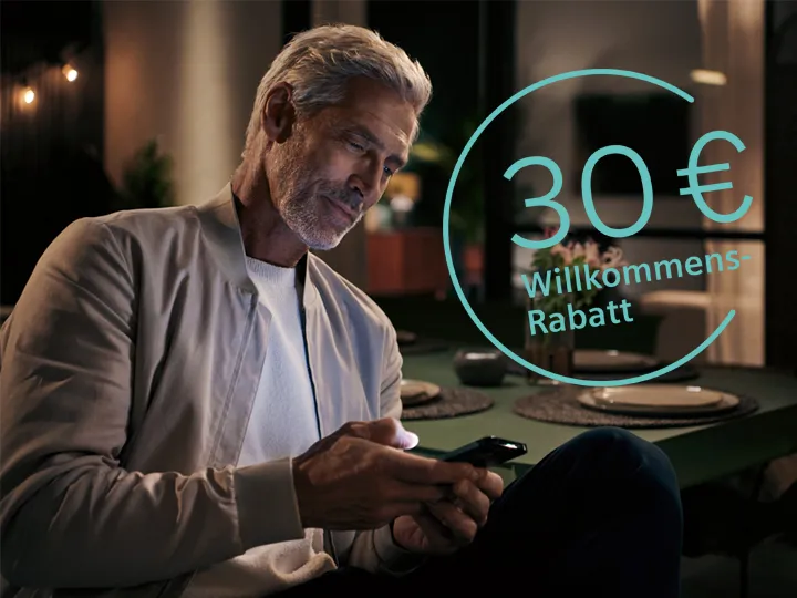 Älterer Mann mit grauem Haar und Bart sitzt in einem dunklen Raum und schaut auf sein Smartphone