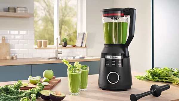Un elegante frullatore nero con un frullato verde all'interno, circondato da ingredienti freschi come cavolo riccio, mele e avocado in una cucina luminosa.