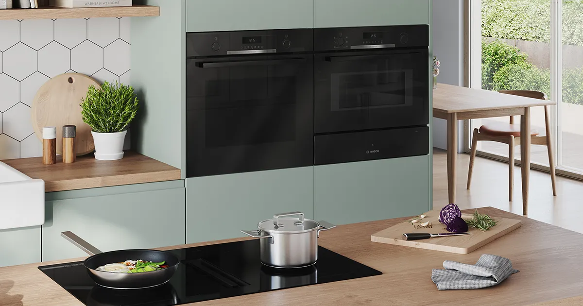 Sodobna kuhinja z mentolovo zelenimi omaricami, črnimi pečicami, ponevjo z zelenjavo in loncem na gladki kuhalni plošči, osvetljena z naravno svetlobo.