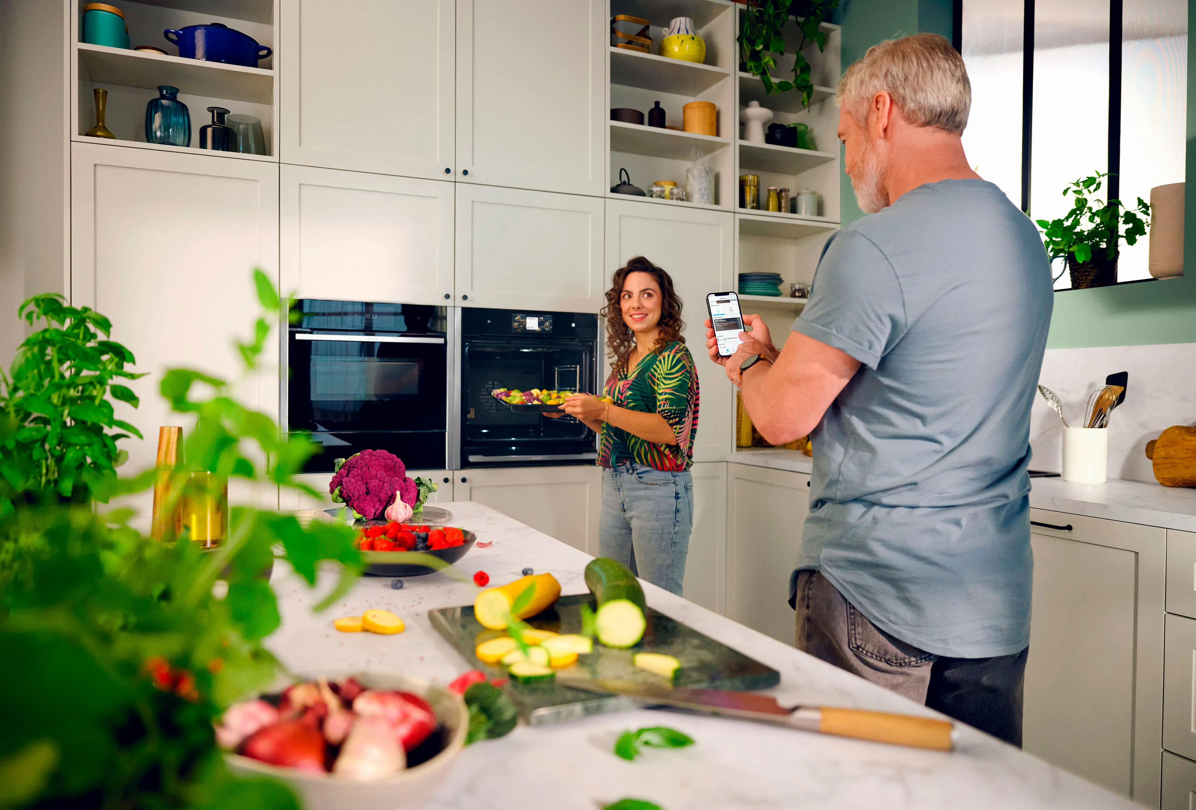 En person lagar mat i ett modernt kök, medan en annan person använder smartphone för att följa recept eller inspelning.
