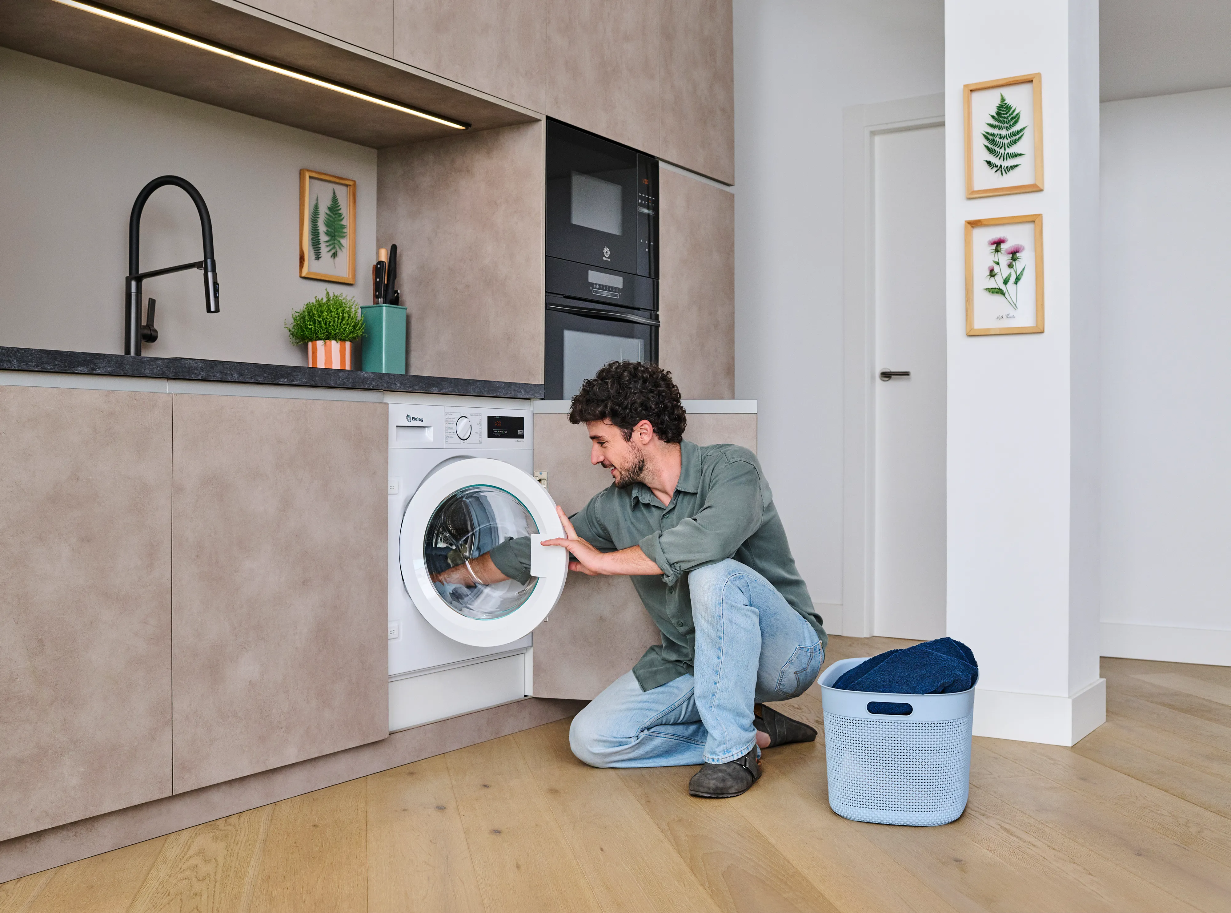 Un hombre se arrodilla para meter la ropa en una lavadora en una cocina moderna, con una cesta de la ropa sucia cerca.