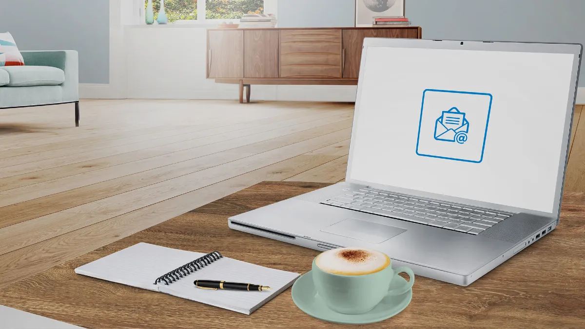 Laptop, notebook, pen, coffee cup on wooden floor with furniture in background.