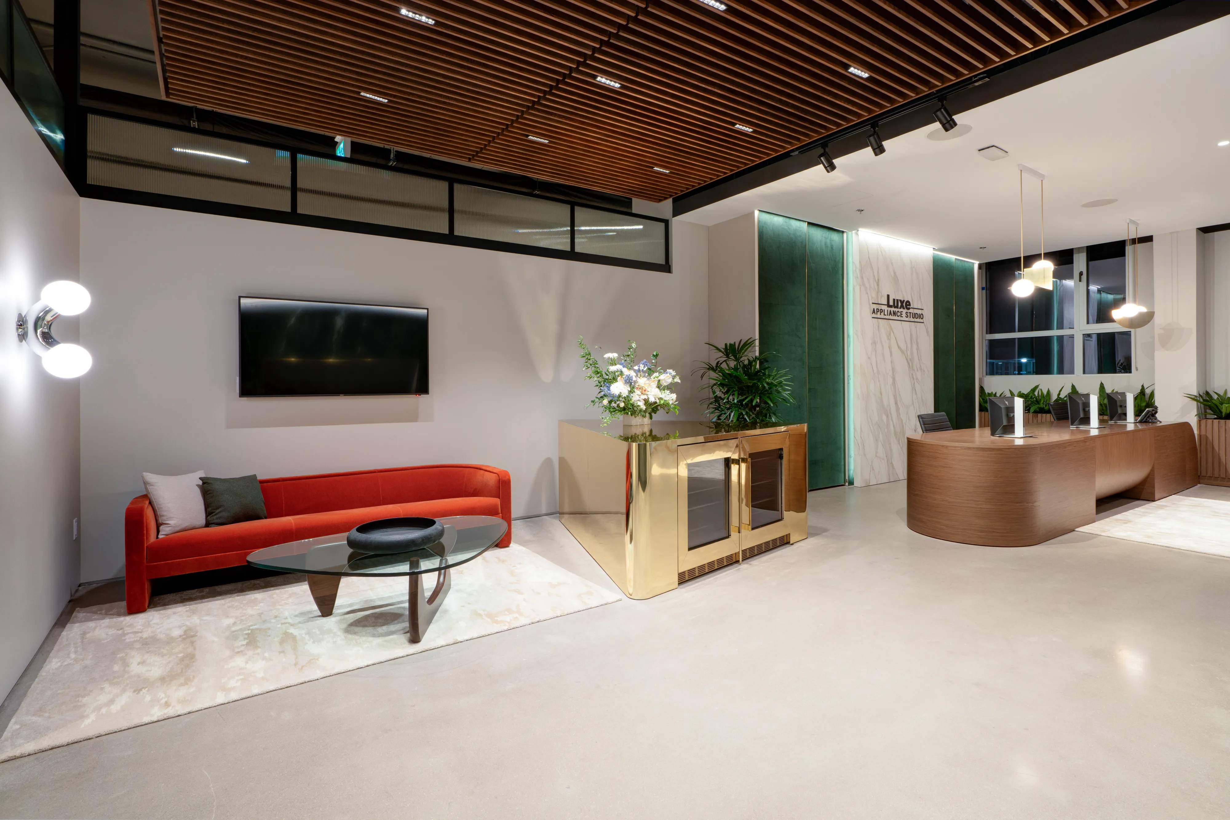 Hall d’entrée d’un bureau moderne avec canapé rouge, table basse en verre, téléviseur, plafond en panneaux de bois et comptoir d’accueil incurvé avec luminaires suspendus.