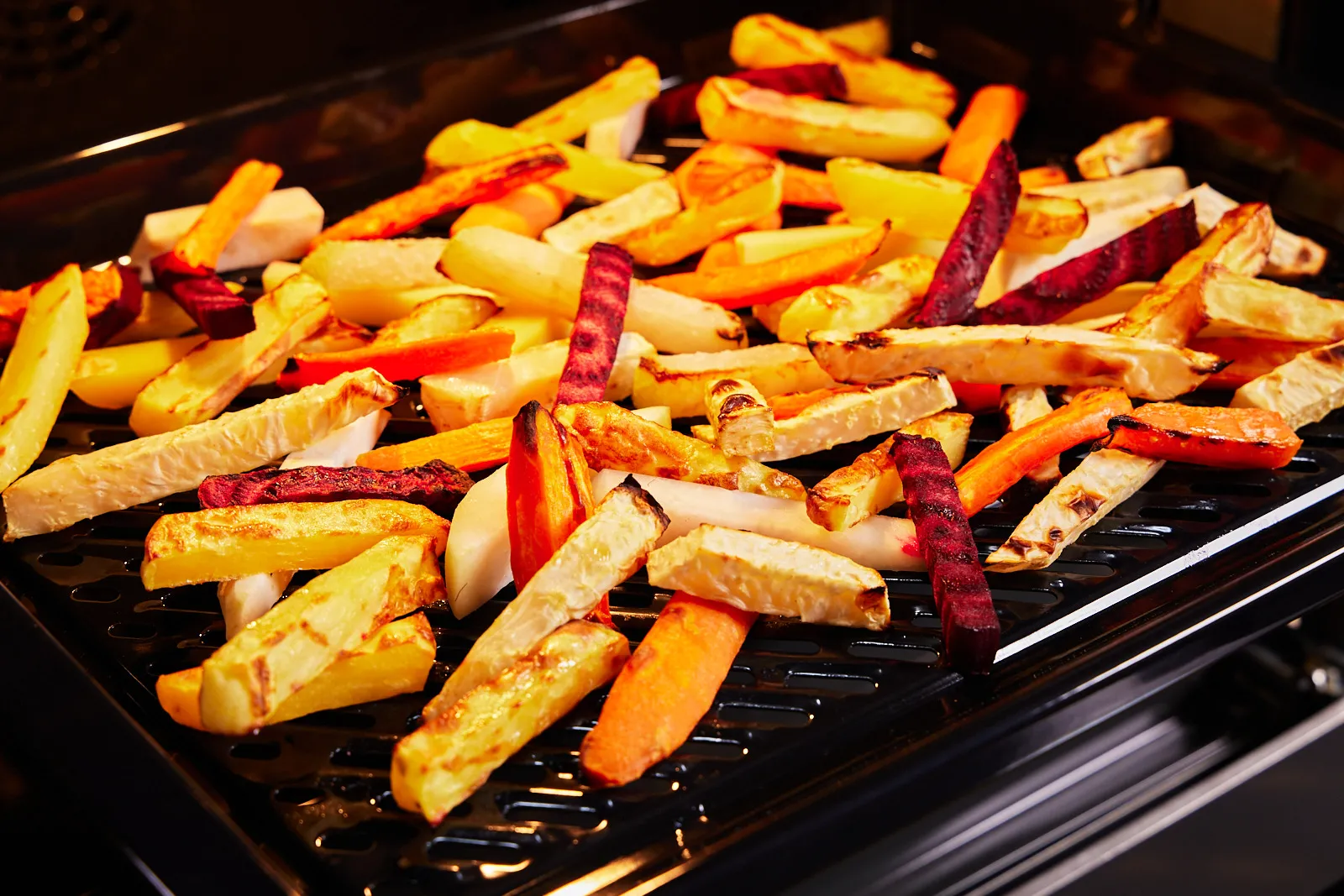 Geröstete, bunte Gemüsestäbchen, darunter Karotten, Rote Bete und Pastinaken, auf einem schwarzen Grillblech.