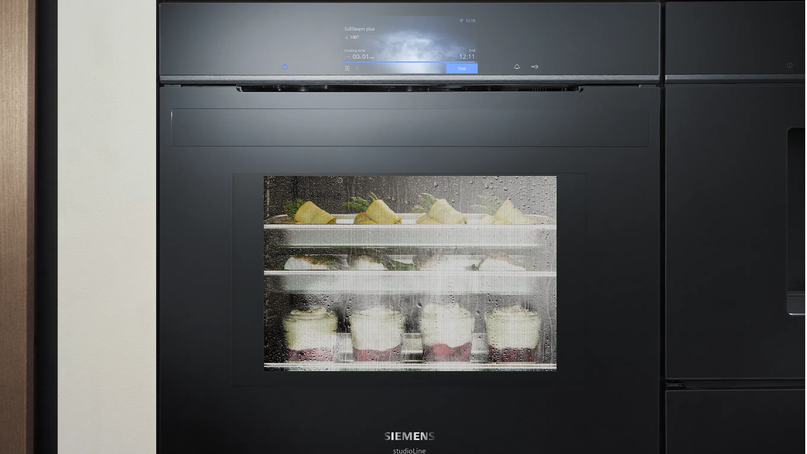 A black Siemens studioLine steam oven is shown with a steamy glass door, through which desserts and other foods can be seen cooking on multiple racks.