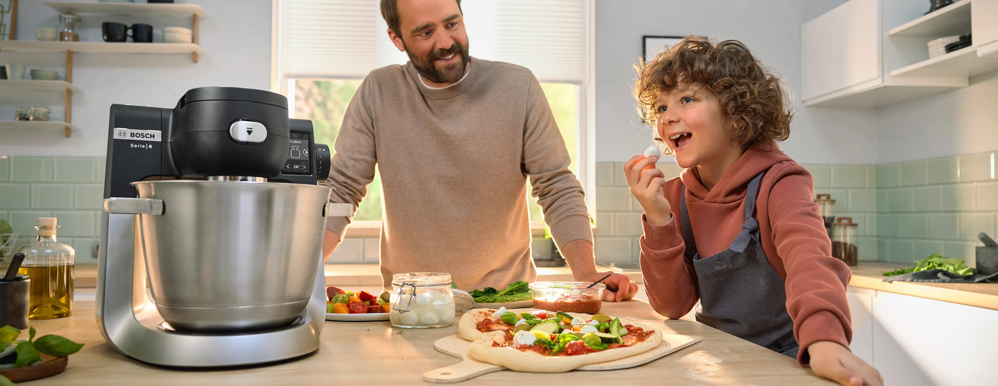 A Bosch kitchen mixer on a countertop with fresh vegetables and pizza ingredients, family cooking together.