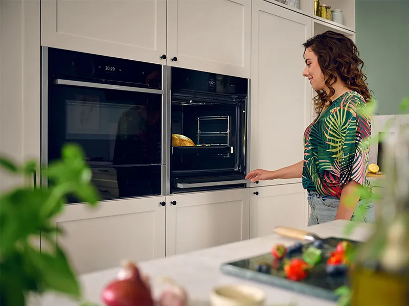 Woman in colorful shirt opening oven in modern kitchen with fresh produce on counter.