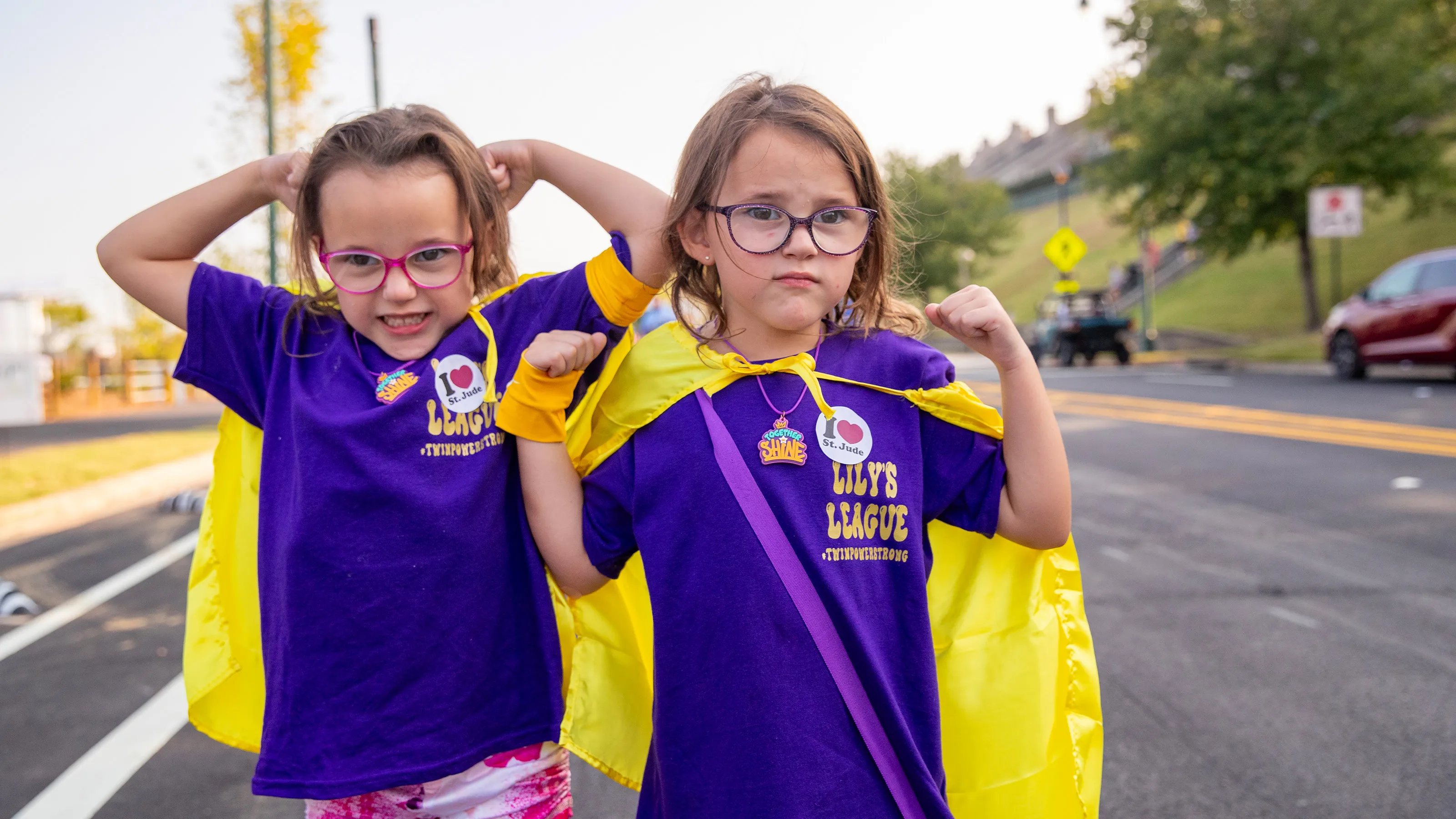 Two children in purple shirts and capes posing confidently in the street.