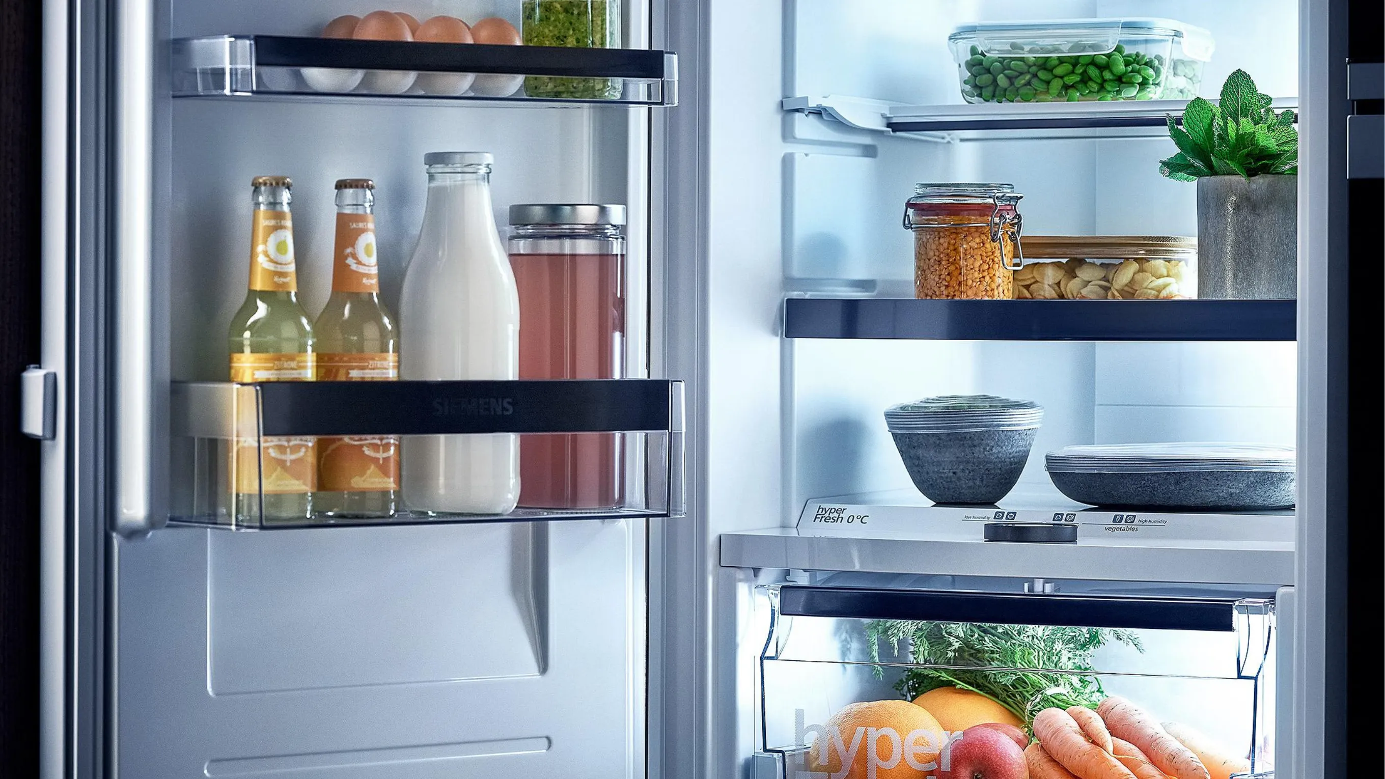 Brightly lit open fridge with organized food and fresh produce, next to a built-in Siemens oven.