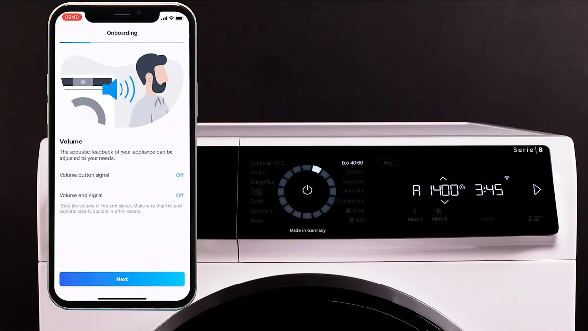 Smartphone visar Home Connect appen, tvättmaskin med digital display, moderna apparater, teknikintegration.