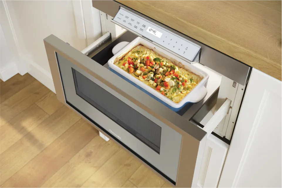 A microwave drawer with a casserole dish filled with vegetable mix is open on a wooden countertop in a modern kitchen.