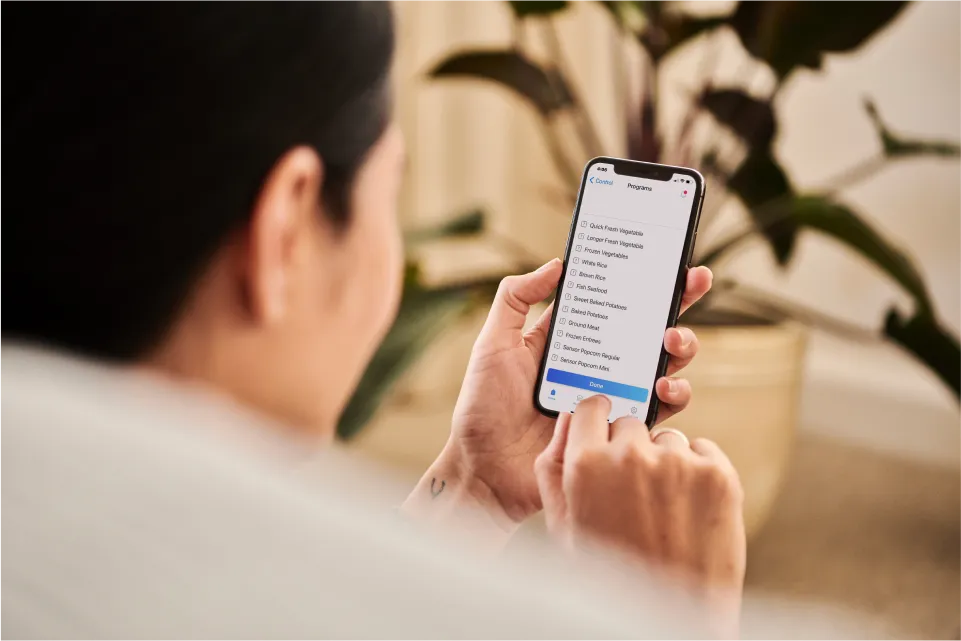 A person holds a smartphone displaying a menu of vegetable options and settings, surrounded by indoor plants.