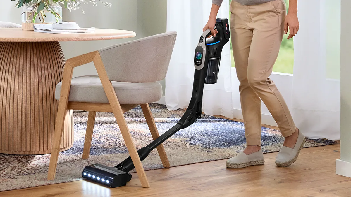 Eine Frau saugt mit einem kabellosen Unlimited Akku-Staubsauger von Bosch unter einem Stuhl am Esstisch.