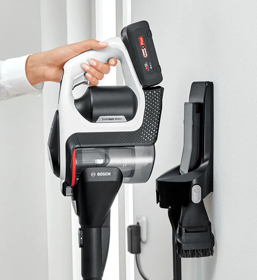 A hand holds the white and black Bosch cordless vacuum cleaner, with its removable battery visible, next to a wall-mounted charging station with an attached brush accessory.