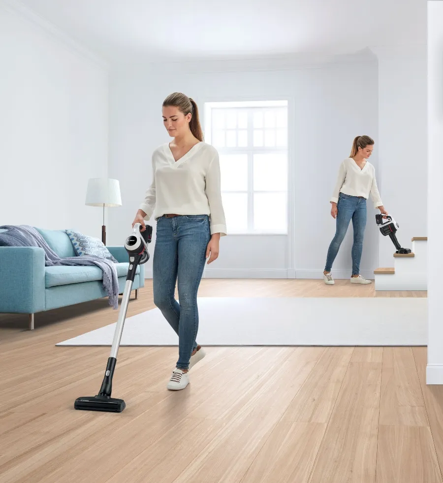 A woman uses a Bosch cordless stick vacuum cleaner to clean a wooden floor, and in a smaller inset, she cleans stairs with the same vacuum, highlighting its versatility.