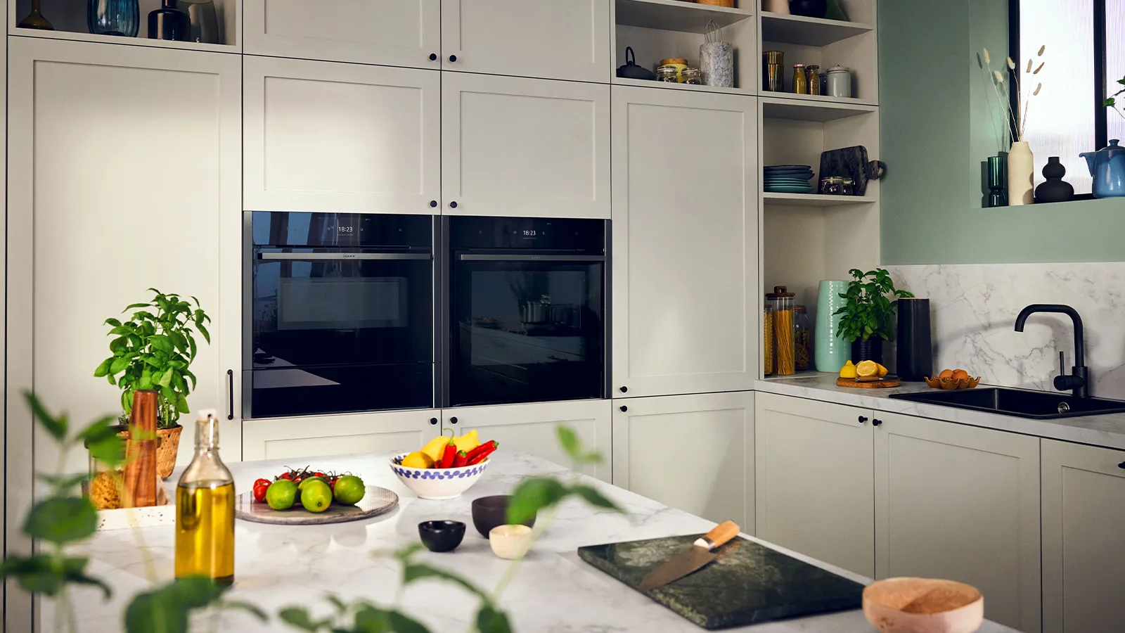 Modern kitchen with white cabinets, built-in ovens, fresh produce, and potted plants.