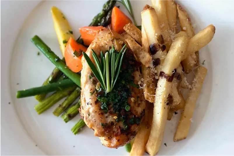 Grilled chicken topped with herbs, served with sautéed vegetables and crispy French fries on a white plate.