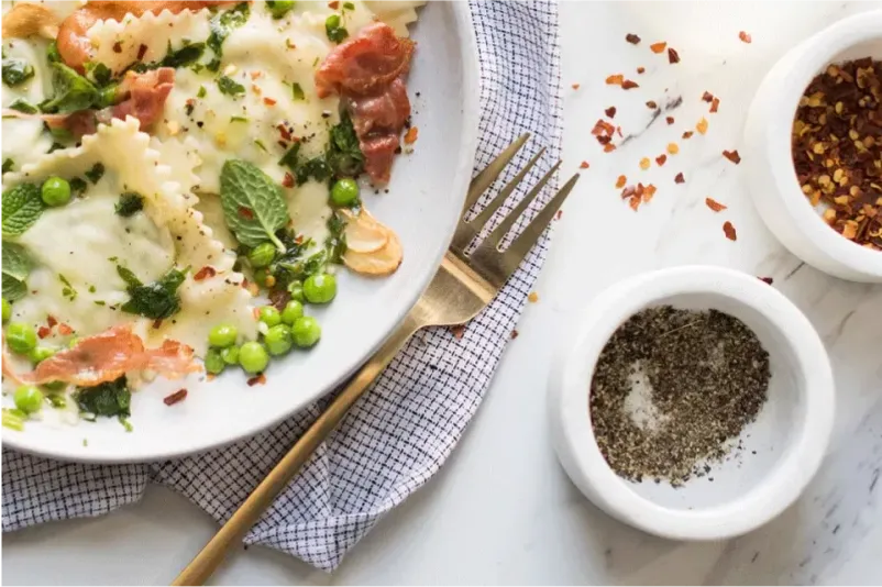 A plate of creamy ravioli topped with ham, peas, and mint, accompanied by salt and pepper pots and red pepper flakes.