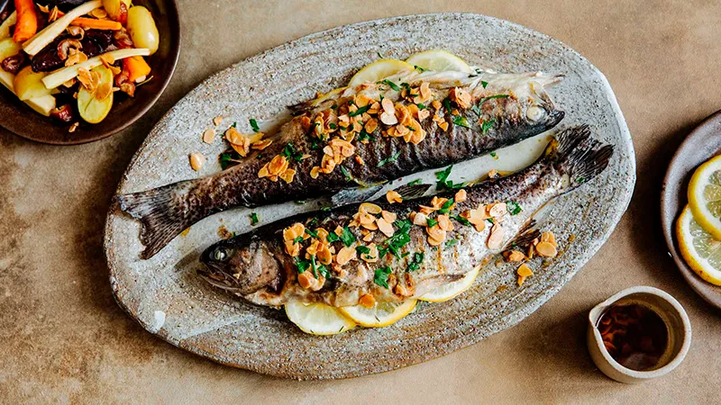 Gegrillte ganze Fisch mit Gemüse, Kräutern und Zitronenscheiben auf einer Steinplatte.
