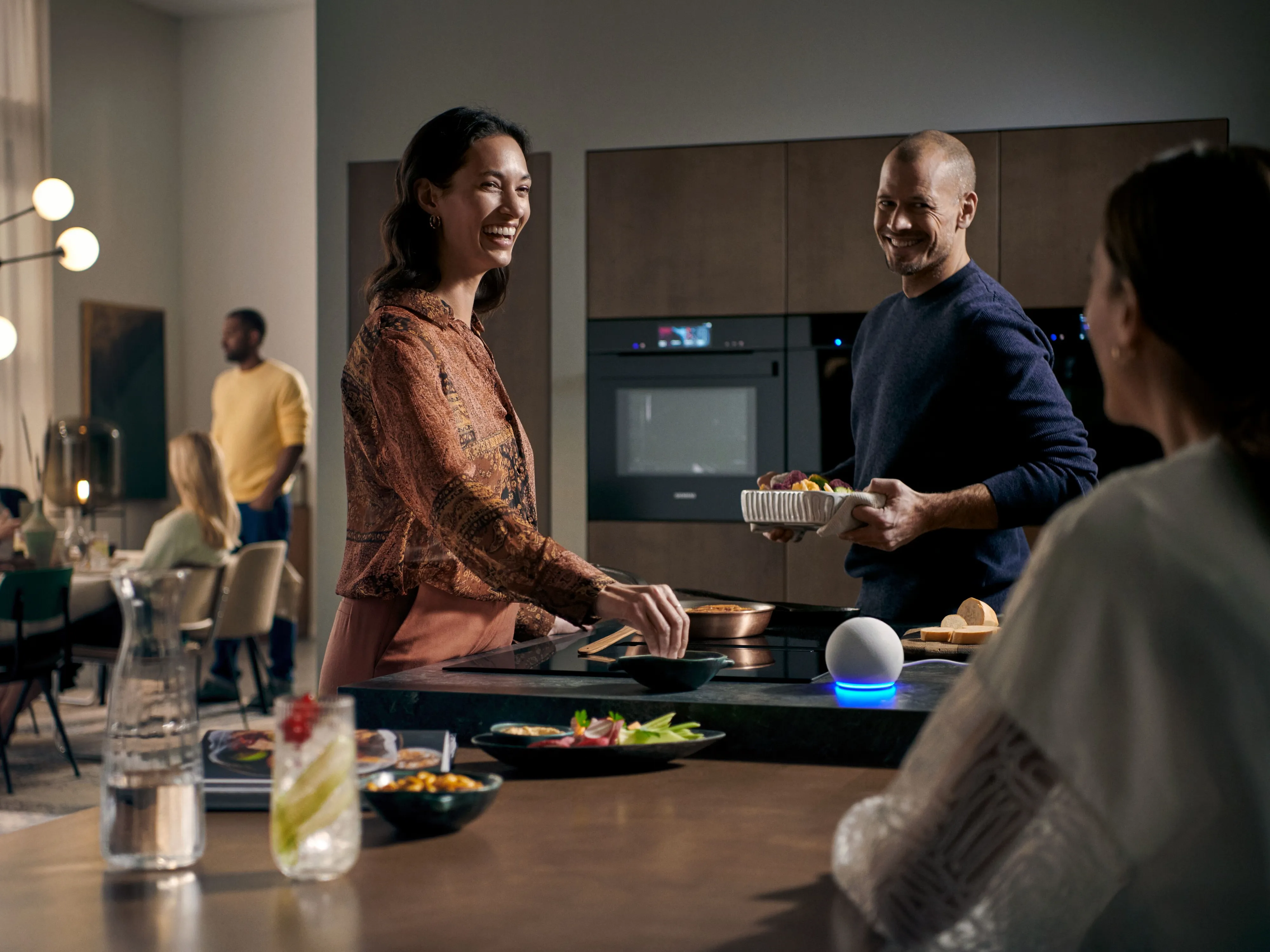 Glade venner forbereder middag i et moderne køkken. De hygger sig og snakker ved køkkenøen med gæster i baggrunden.