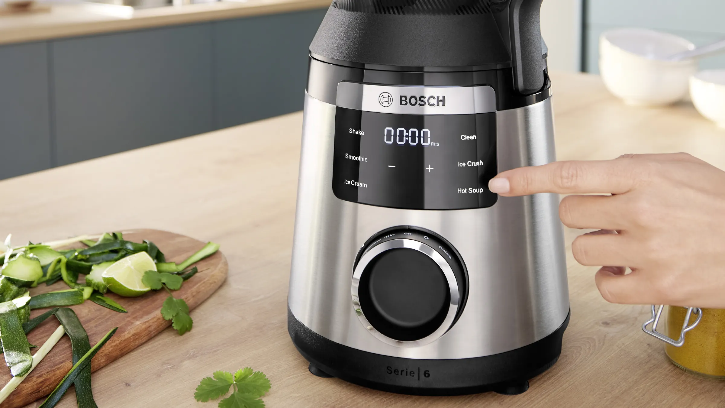Prim plan cu o mână care indică afişajul digital al blenderului VitaPower Serie 6 aflat pe un blat din bucătărie, lângă ingrediente verzi, precum limete şi frunze.