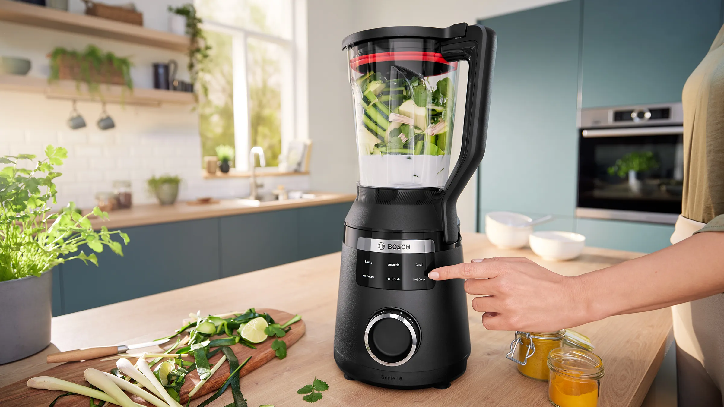 O mână care indică spre afişajul digital al blenderului VitaPower Serie 6, al cărui vas este plin cu legume verzi şi este aşezat pe un blat de bucătărie, alături produse verzi, precum limete, lemongrass şi frunze de coriandru.