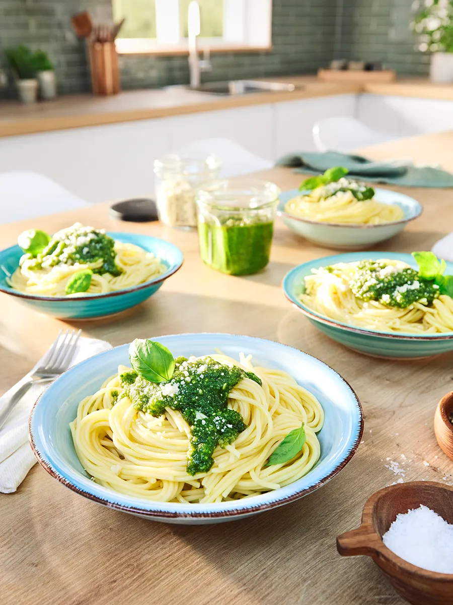 Vier kommen pasta met groene pesto, gerangschikt op een keukentafel met een pot pesto in het midden.
