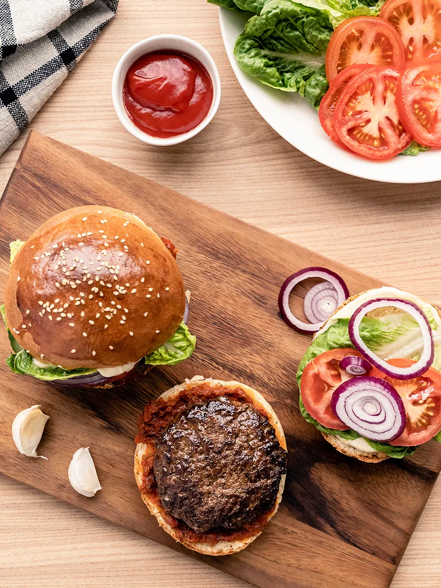 Vogelperspectief van een burgerpasteitje op een briochebroodje naast schijven tomaten, uien en sla op een houten oppervlak.