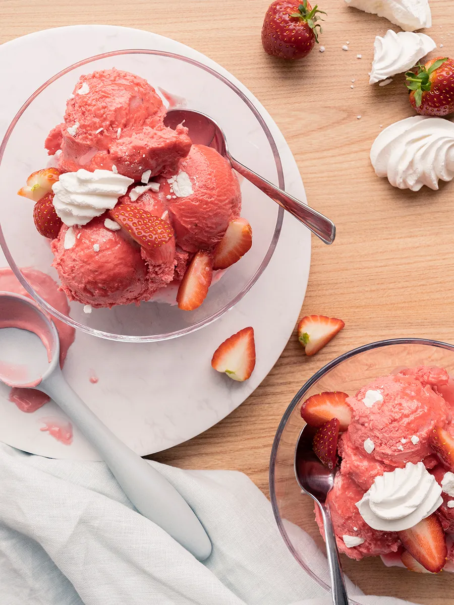 2 glazen dessertkommen met aardbeienijs, gegarneerd met aardbeien en meringue op een houten oppervlak.