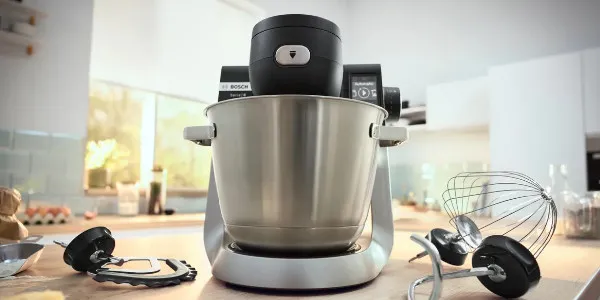 Un moderno mixer da cucina Bosch con ciotola, fruste e accessori in acciaio inox, posizionato su un piano di lavoro in legno in una luminosa cucina.