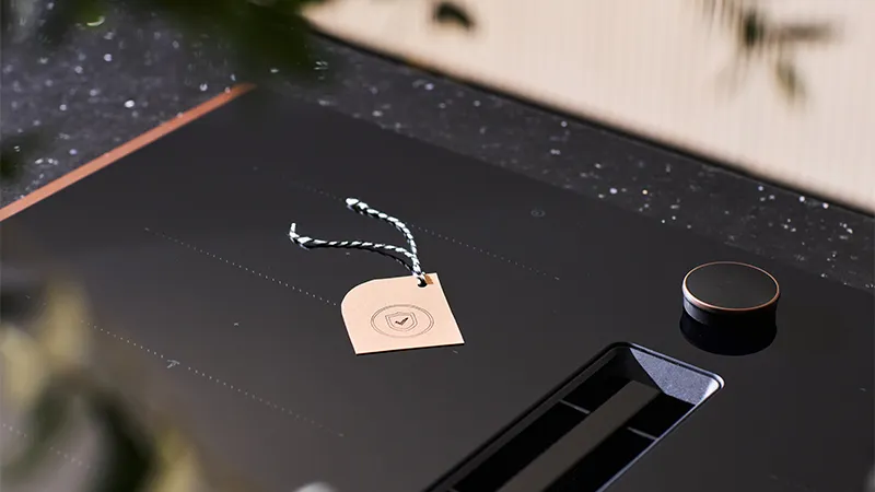 alt-Kaasaegsel mustal induktsioonpliidil on beež silt linnukesega, mis näitab, et pliidiplaat on garantii all. See on paigutatud kergelt laigulisele tööpinnale ja roheline lehestik raamib stseeni õrnalt.