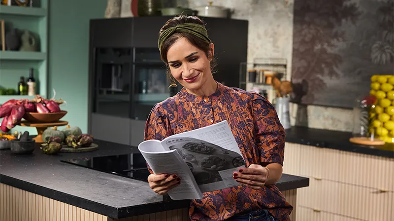 alt-A NEFF customer stands in a stylish kitchen, reading a user manual, surrounded by colorful ingredients and kitchenware.