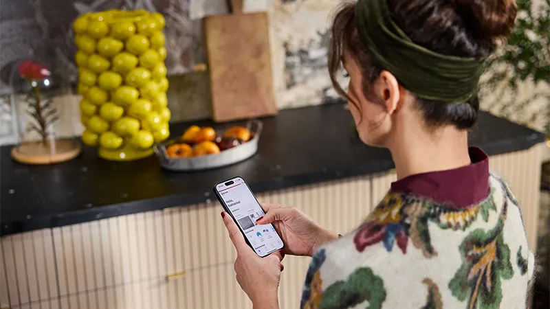 alt-A person in a cosy kitchen, surrounded by a bowl of oranges and a vibrant vase of lemons, navigates to NEFF Self Help content using a smartphone.