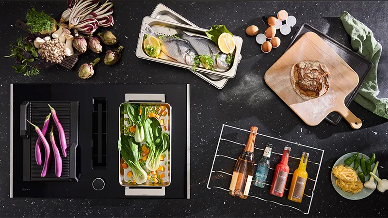 alt-A modern kitchen setup with NEFF cooking accessories, fresh vegetables, and colorful drinks arranged around a sleek cooking surface.
