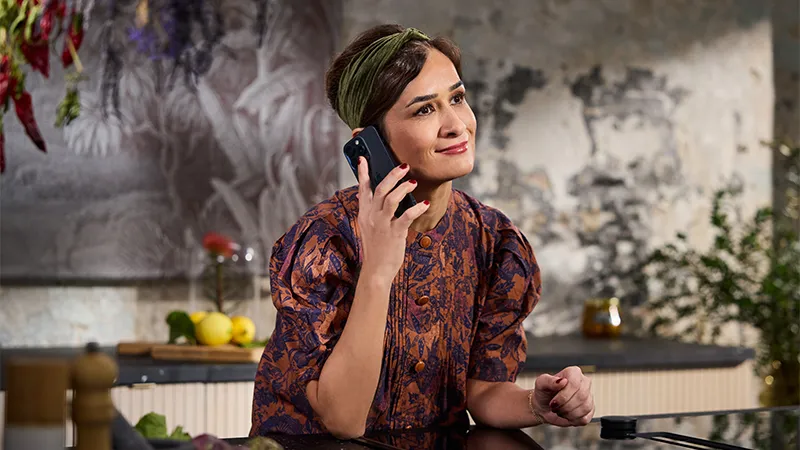 alt-A person in a patterned shirt and headband is sitting at a table, using a smartphone to call NEFF customer service, surrounded by a rustic kitchen setting.