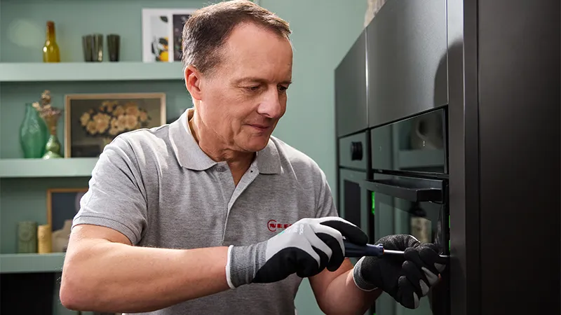 alt-A NEFF technician wearing gloves works on a modern oven, using a screwdriver against a backdrop of home decor and green walls.