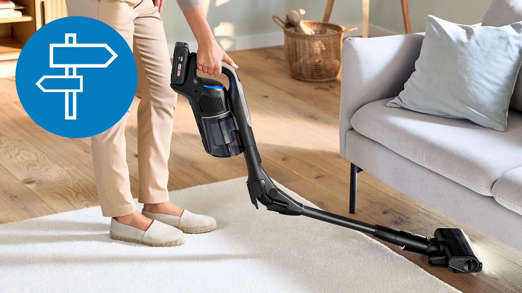 A person using a cordless vacuum cleaner on a white rug, with a directional sign icon in the corner.