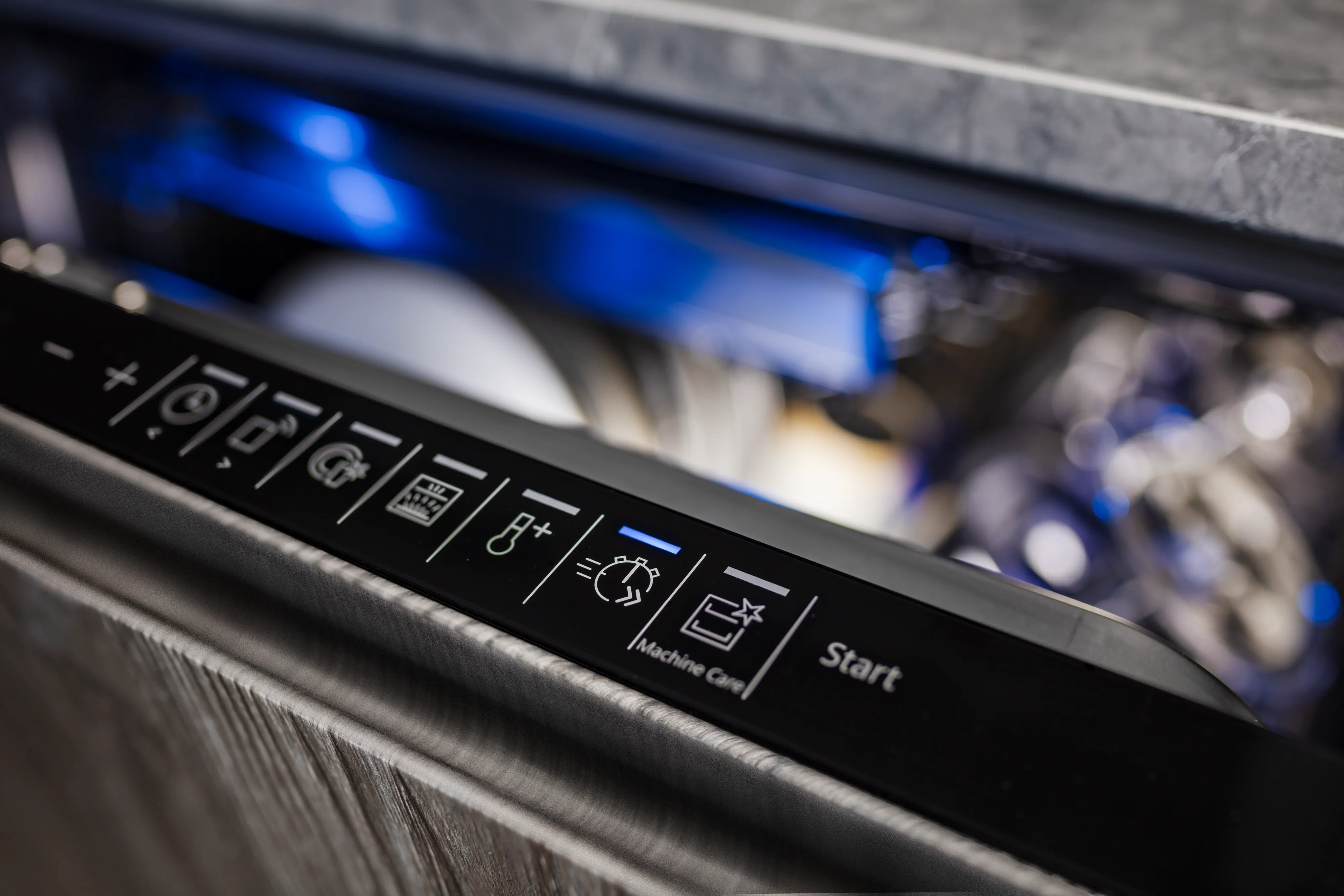 A close-up of the black control panel on an integrated dishwasher. The panel, which sits atop a dark wood-grain door, displays various wash cycle icons, with the timer/delay start symbol illuminated by a blue light. The blurred interior of the dishwasher is visible in the background, also glowing with blue light.