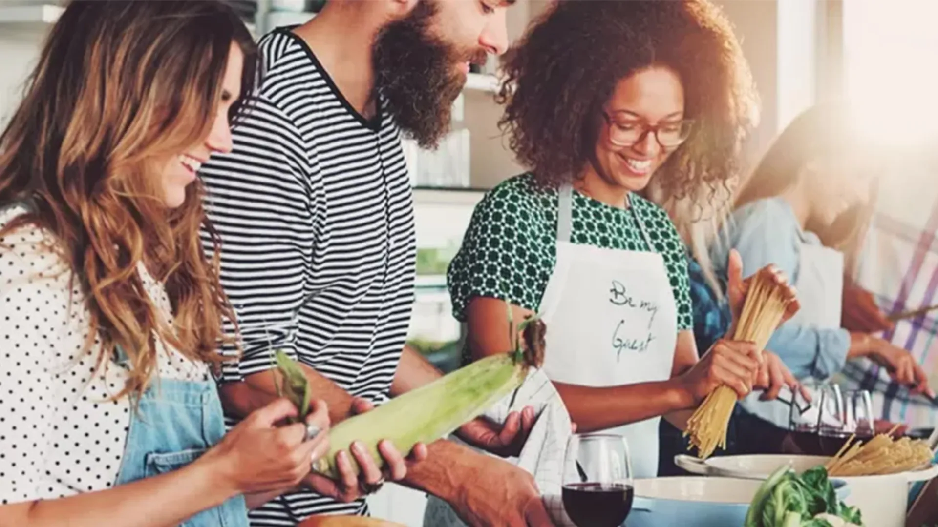 Vier Freunde bereiten gut gelaunt gemeinsam ein Essen in der Küche zu, halten Mais und Pasta in den Händen, während Weingläser auf der Arbeitsplatte bereitstehen.