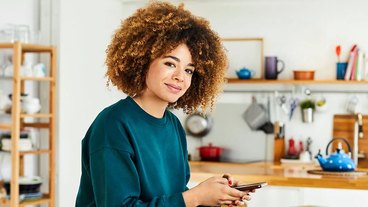 Woman with curly hair in green sweater holding smartphone in kitchen.
