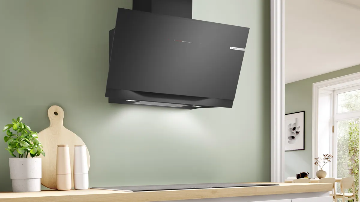 A sleek black Bosch angled extractor hood with integrated lighting is mounted on a sage green kitchen wall above a black induction hob and wooden countertop.