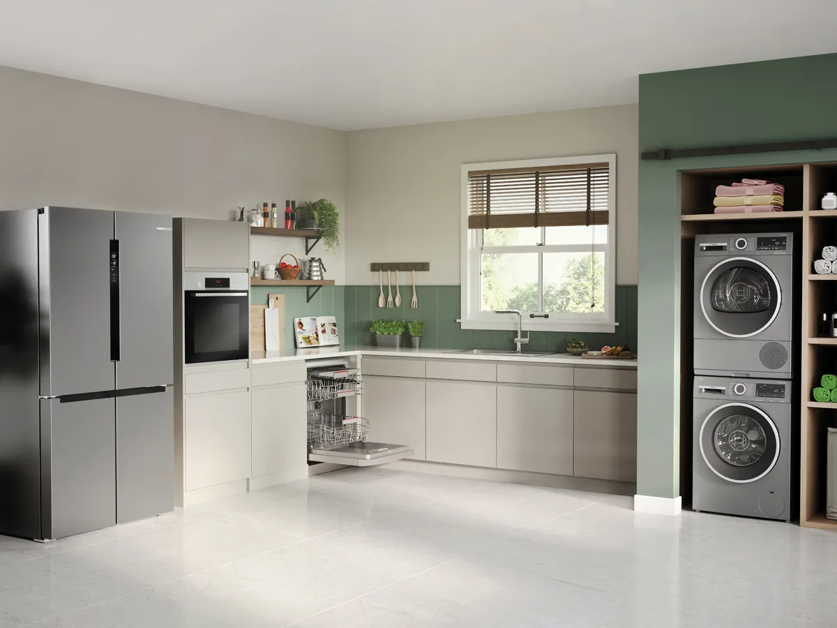 A spacious, modern kitchen and laundry room with light grey and green walls, featuring a stainless steel French door refrigerator, an integrated oven, an open dishwasher, and stacked washer and dryer units.