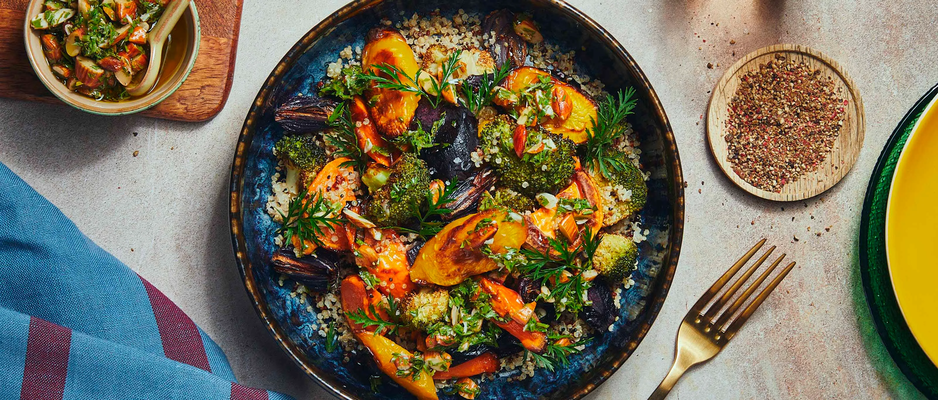 A blue and black ceramic bowl filled with tri colored quinoa which is topped with air fried vegetables is standing in the middle of a table and surrounded by colorful dinner table dishes, like a fork, a napkin, plates and a glas filled with water.