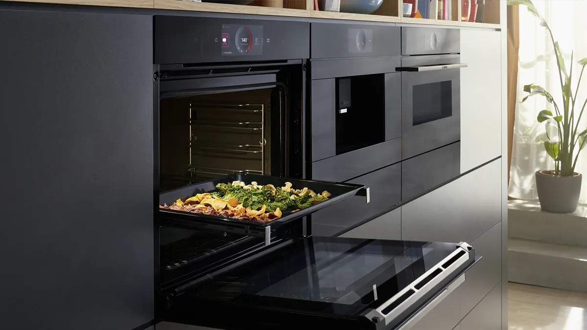 A black Bosch oven with its door open, revealing a baking tray of roasted vegetables and chips being pulled out, integrated into dark cabinetry with other Bosch appliances.